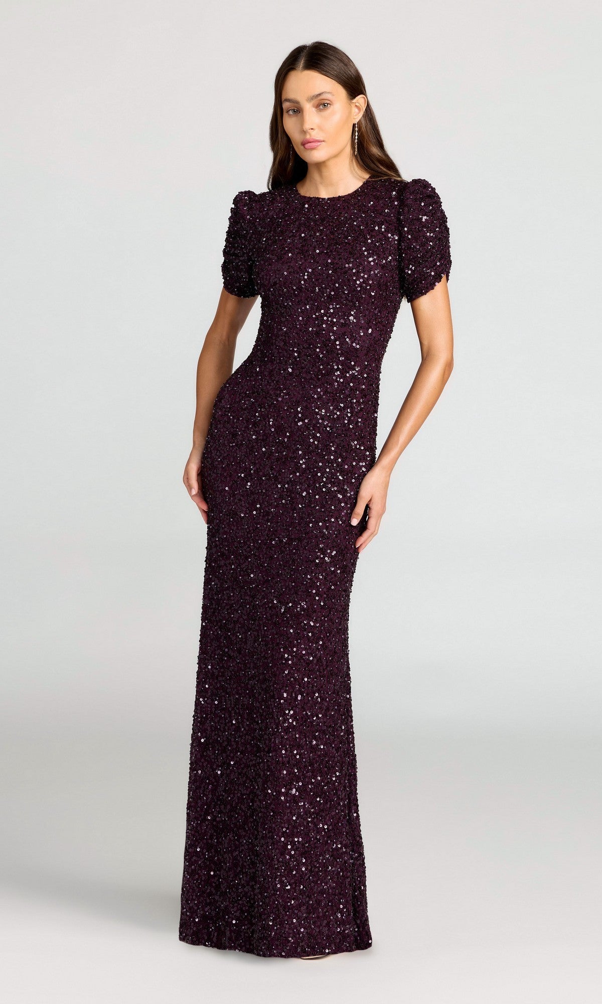 A woman wears the Mac Duggal 24003 Short Sleeve Sequin Formal Dress in Blackberry, featuring puffed sleeves and a floor-length silhouette, set against a plain background.